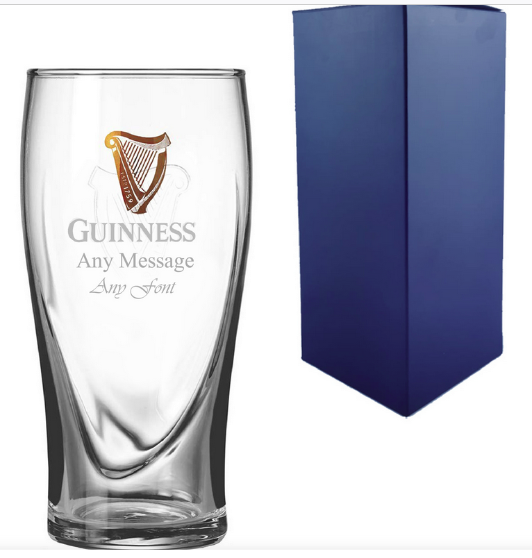 Clear glass Guinness beer pint with engraved harp logo and text next to a blue gift box on a white background